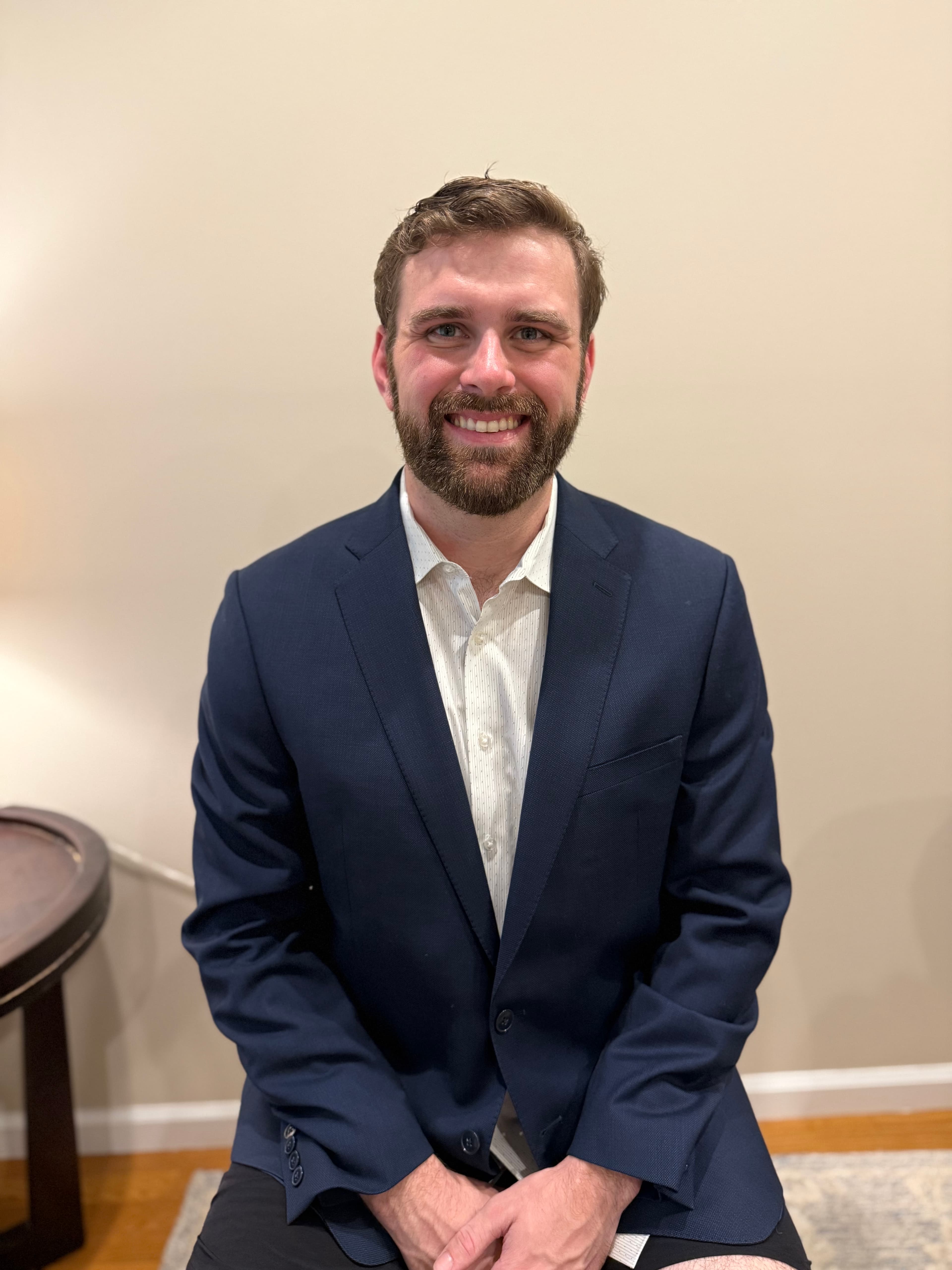Smiling man with a beard wearing a navy blue blazer and white patterned shirt.